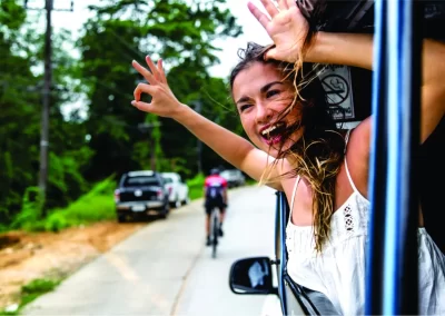 A smiling traveller leaning out of a vehicle window with her arms raised on a countryside road in Puerto Plata